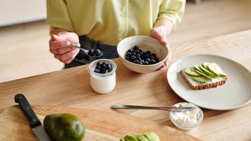 A person prepares a healthy breakfast with yogurt, blueberries, and avocado toast.